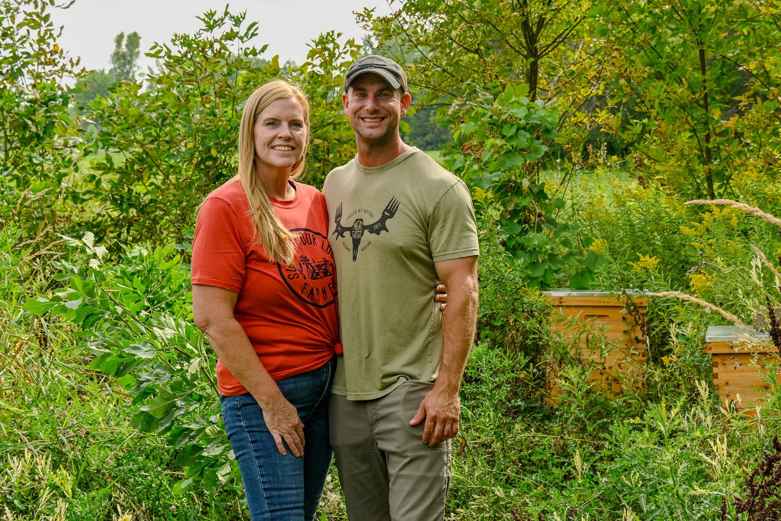 Drew and Sorell standing together in front of green garden growth and beehives