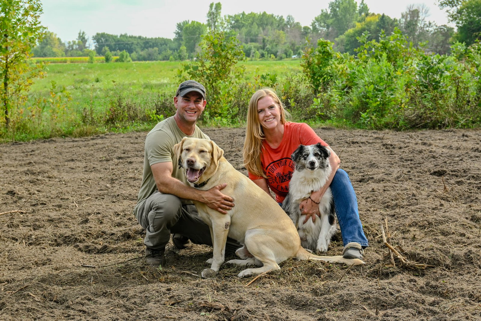 Drew and Sorell kneeling in a freshly worked field with Apollo the yellow Lab and Casey the Mini Australian Shepherd