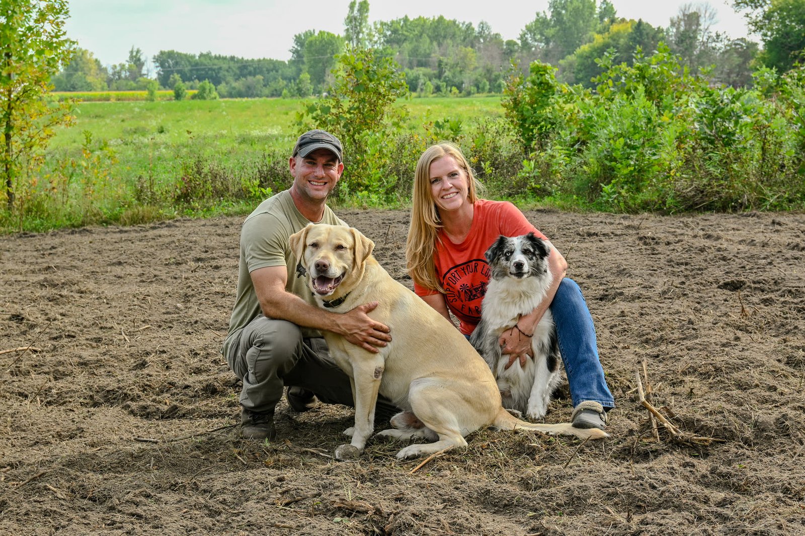Drew and Sorell kneeling in a freshly worked field with Apollo the yellow Lab and Casey the Mini Australian Shepherd